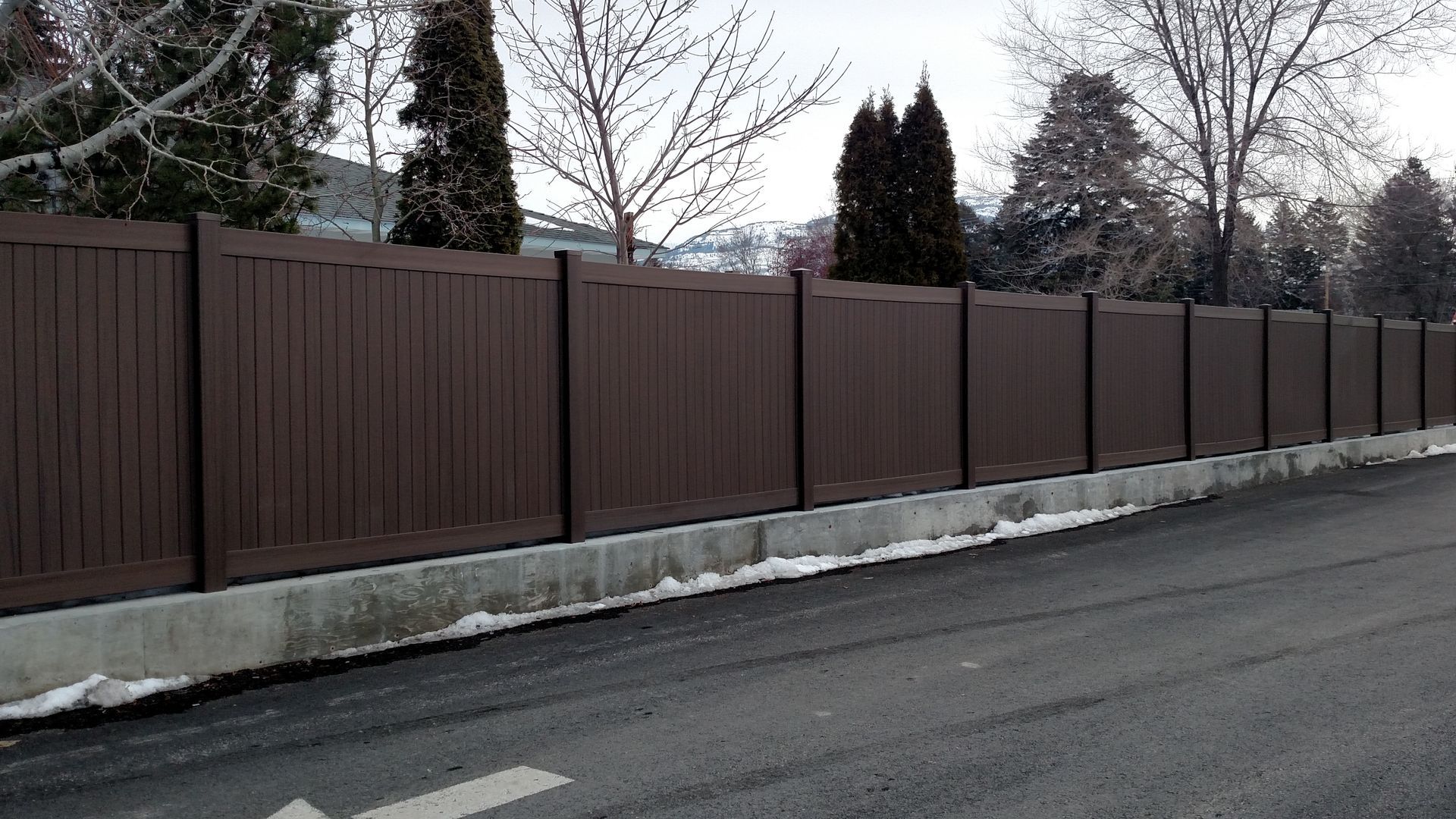 Brown wooden fence lining a snowy roadside with bare trees in the background