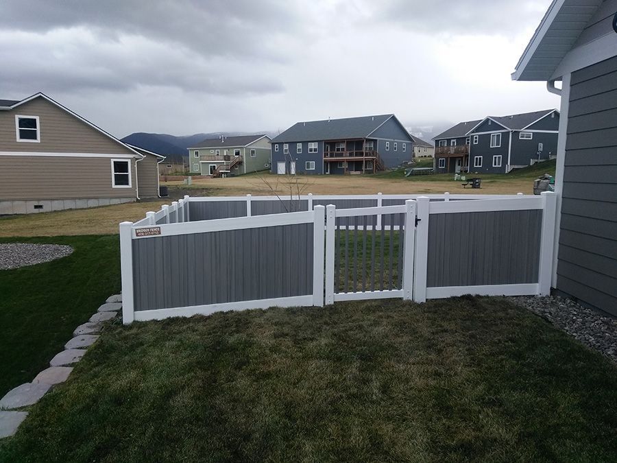 A gray and white fence surrounds a yard with houses in the background.