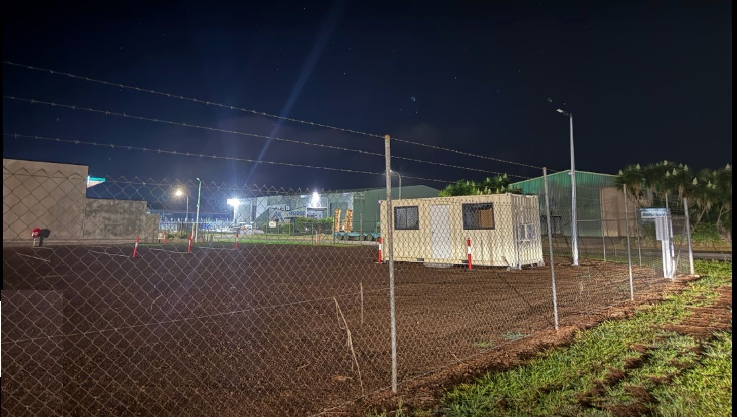 A small, light-colored portable building sits inside a fenced, dirt lot at night under bright overhead security lights Tableland Self Storage In Atherton, QLD