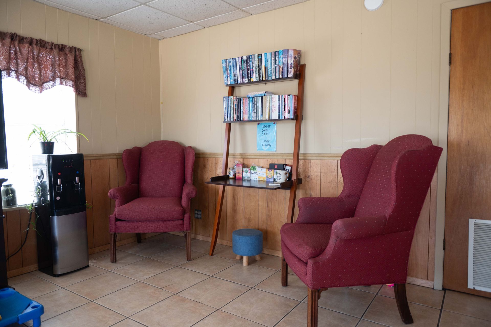 Cozy living room with two burgundy chairs, bookshelf, small fridge, and tiled floor.
