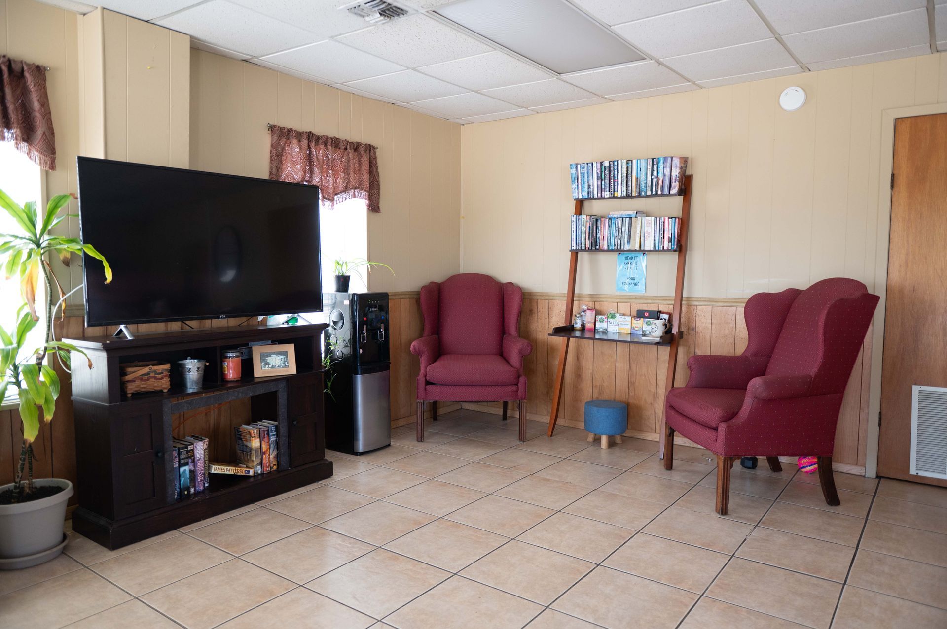 Living room with TV, two maroon chairs, tiled floor, and a small bookshelf against beige walls
