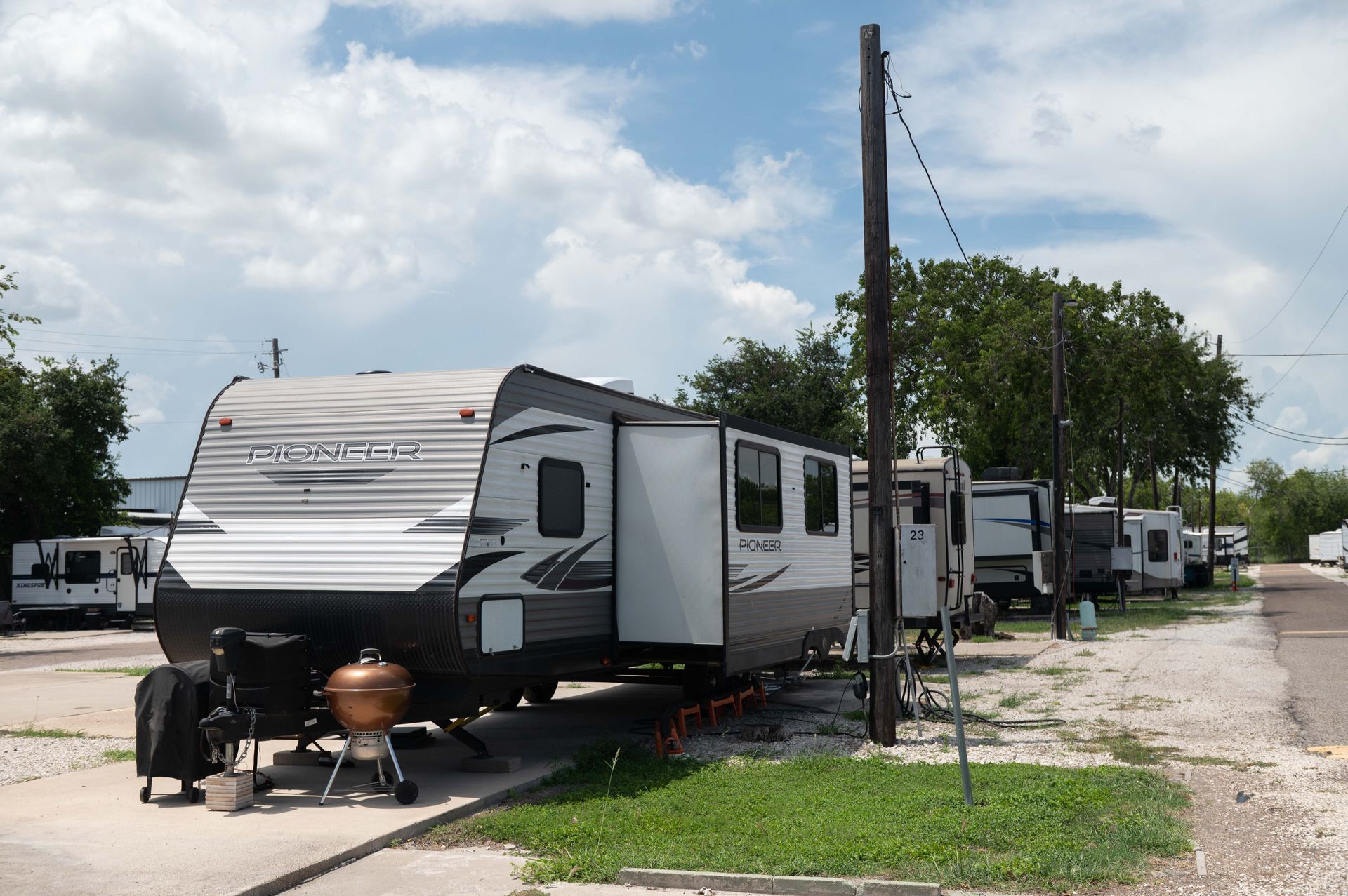 White travel trailer parked at a campsite with awning, chairs, and a propane grill under a cloudy sky