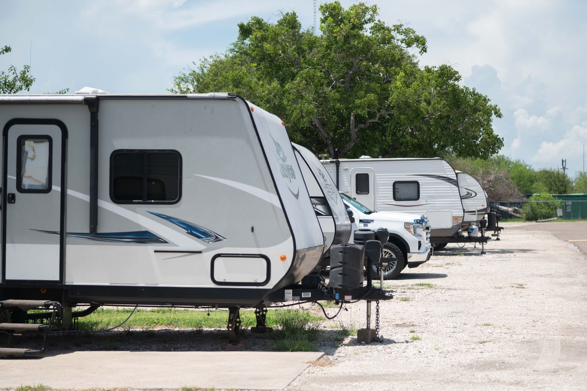 Row of white RV trailers parked in a sunny campground with trees and gravel lot