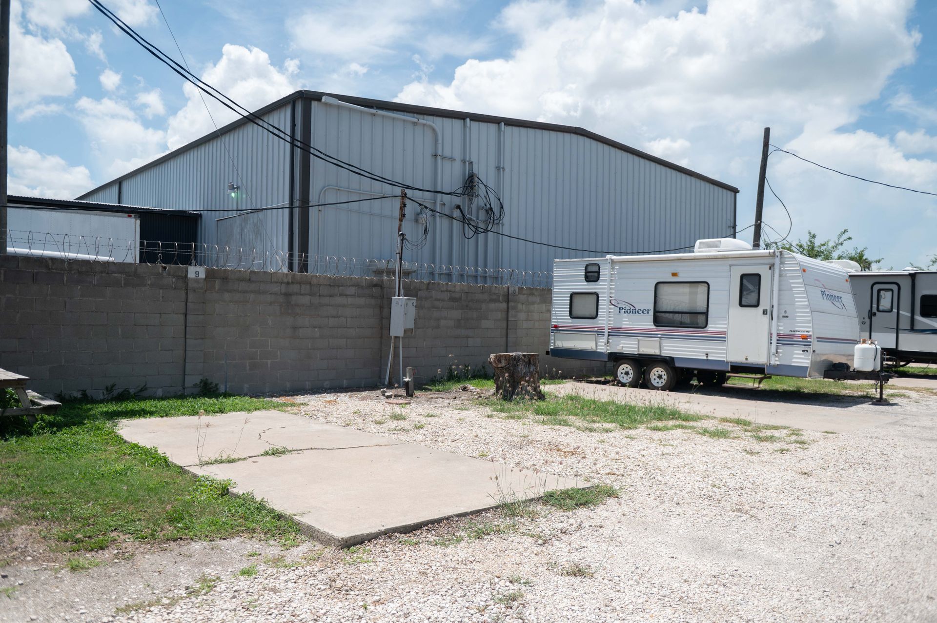 Industrial warehouse yard with gravel lot, concrete pad, and a parked white RV under cloudy sky