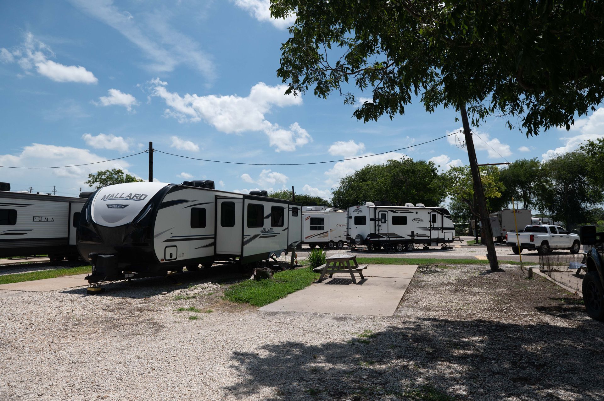 RVs parked at a campsite with picnic tables, gravel ground, and a blue sky with clouds.