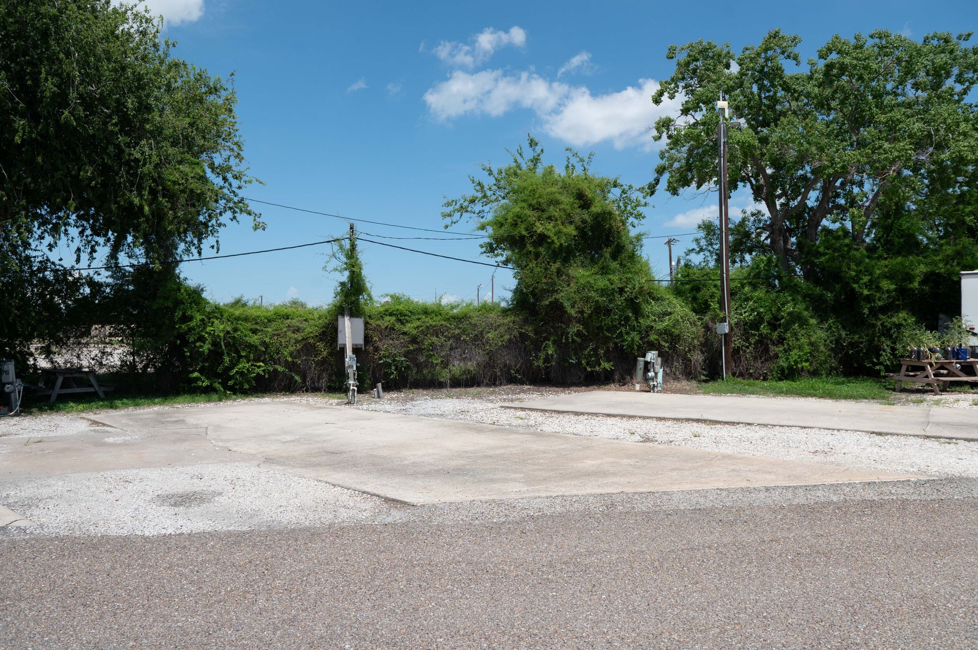 Empty concrete lot beside a dirt road, bordered by trees and a blue sky with clouds.