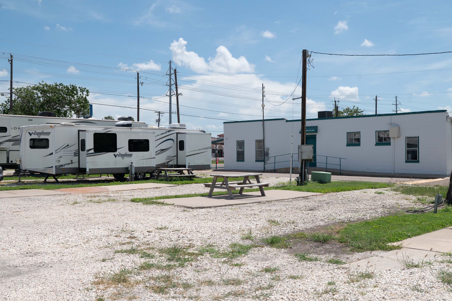 RV park with white trailers, a small building, and a picnic table on gravel under a blue sky