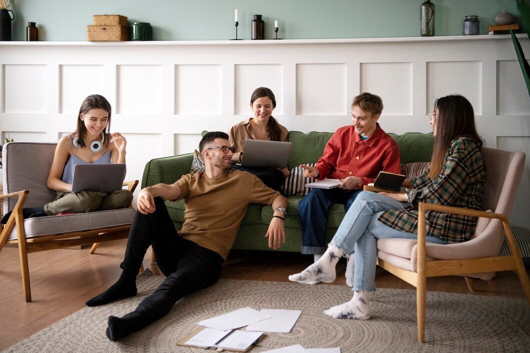 Five people relaxing in a living room, using laptops and phones on a green sofa and chairs.