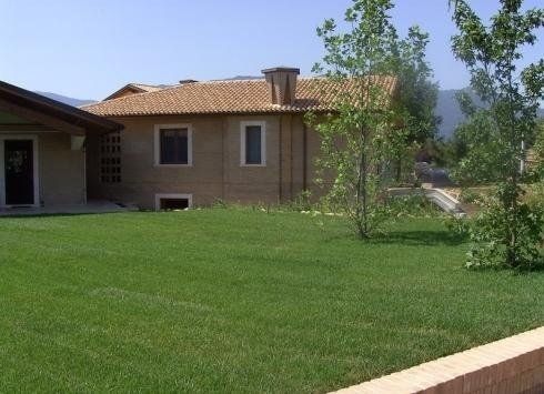 Dietro un prato verde ben curato con piccoli alberi si trova una casa con il tetto in terracotta, le pareti color cuoio e un camino.
