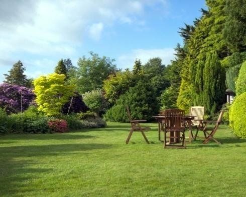 Un tavolo e delle sedie in legno sono sistemati su un prato verde in un lussureggiante giardino ricco di alberi di vario tipo e arbusti in fiore.