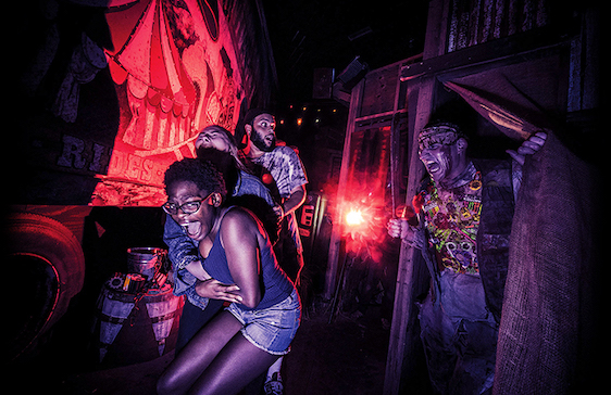 People in a haunted house scream as a zombie reaches towards them. Red light, dark setting.