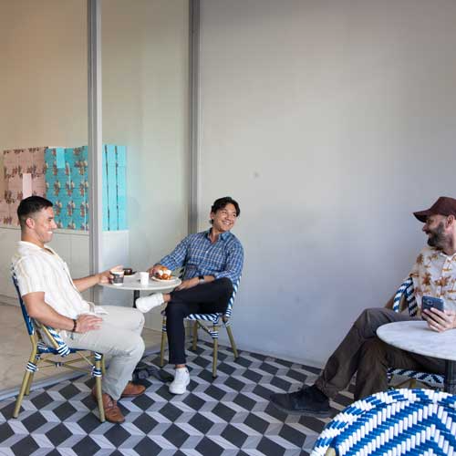 Three men sit at small tables in a cafe, laughing. Black and white checkered floor, colorful chairs.