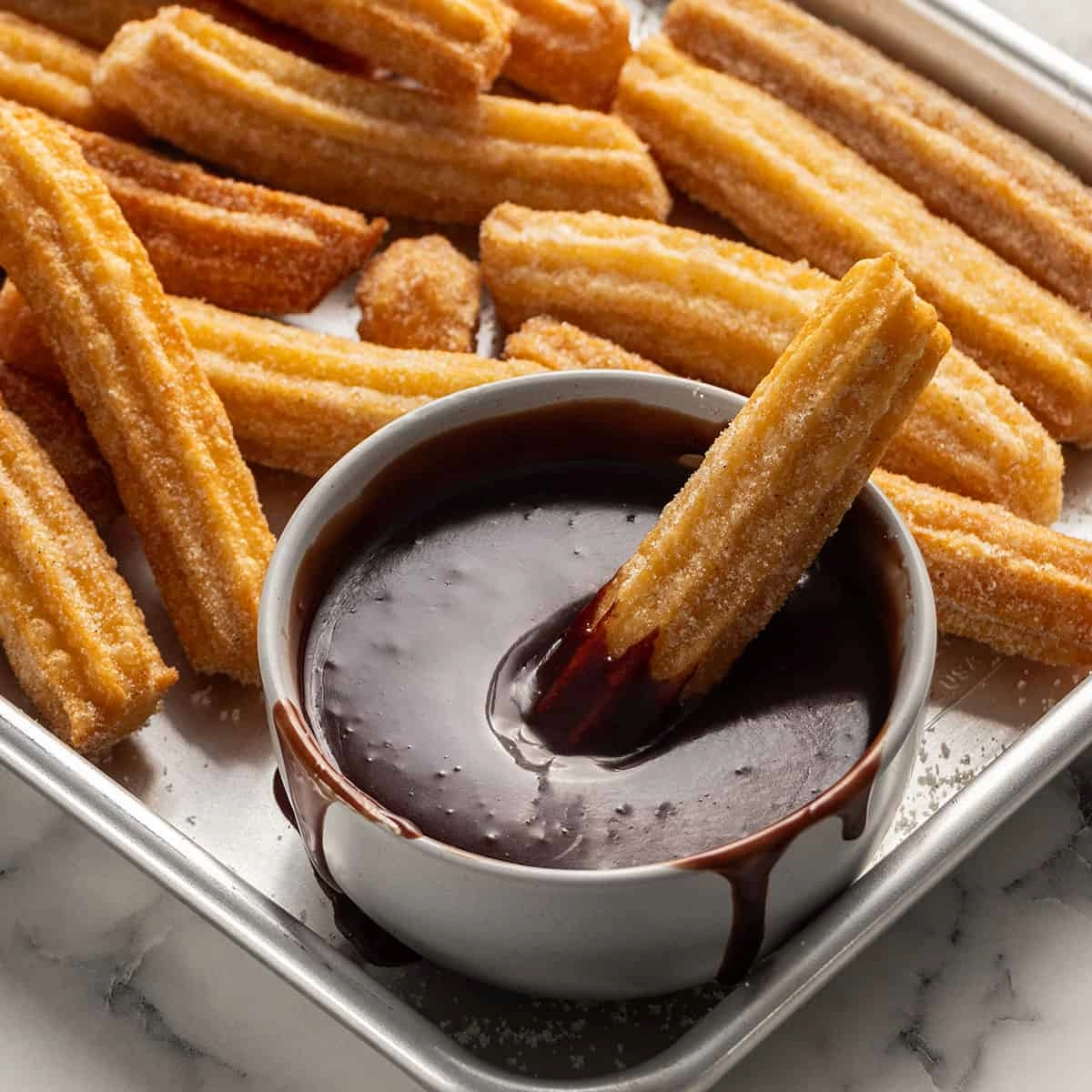 Churros on a tray, one dipped in chocolate sauce. Golden-brown, sweet, fried pastry sticks.
