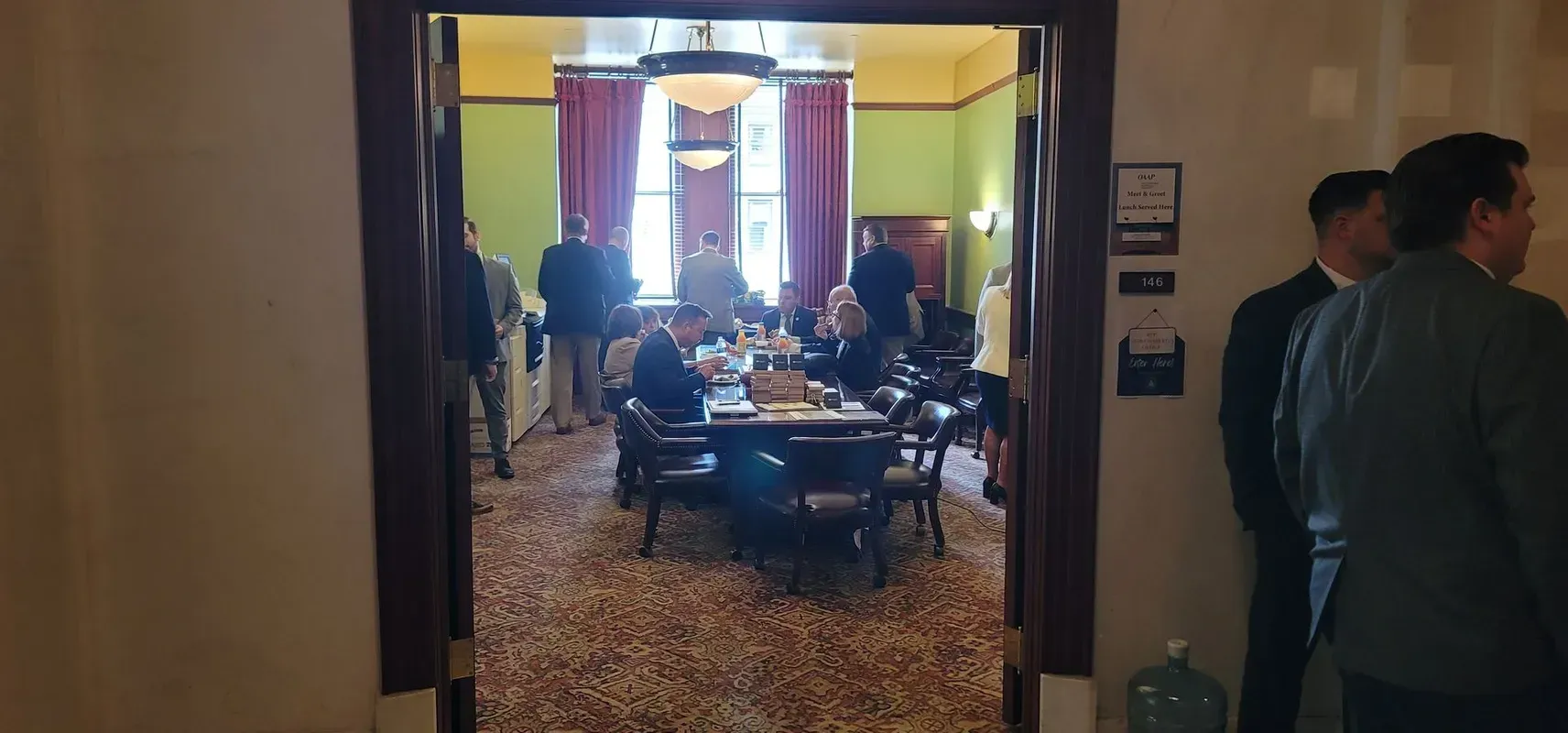 People in a room with a table, seen through a doorway. The room has green walls and patterned carpet.