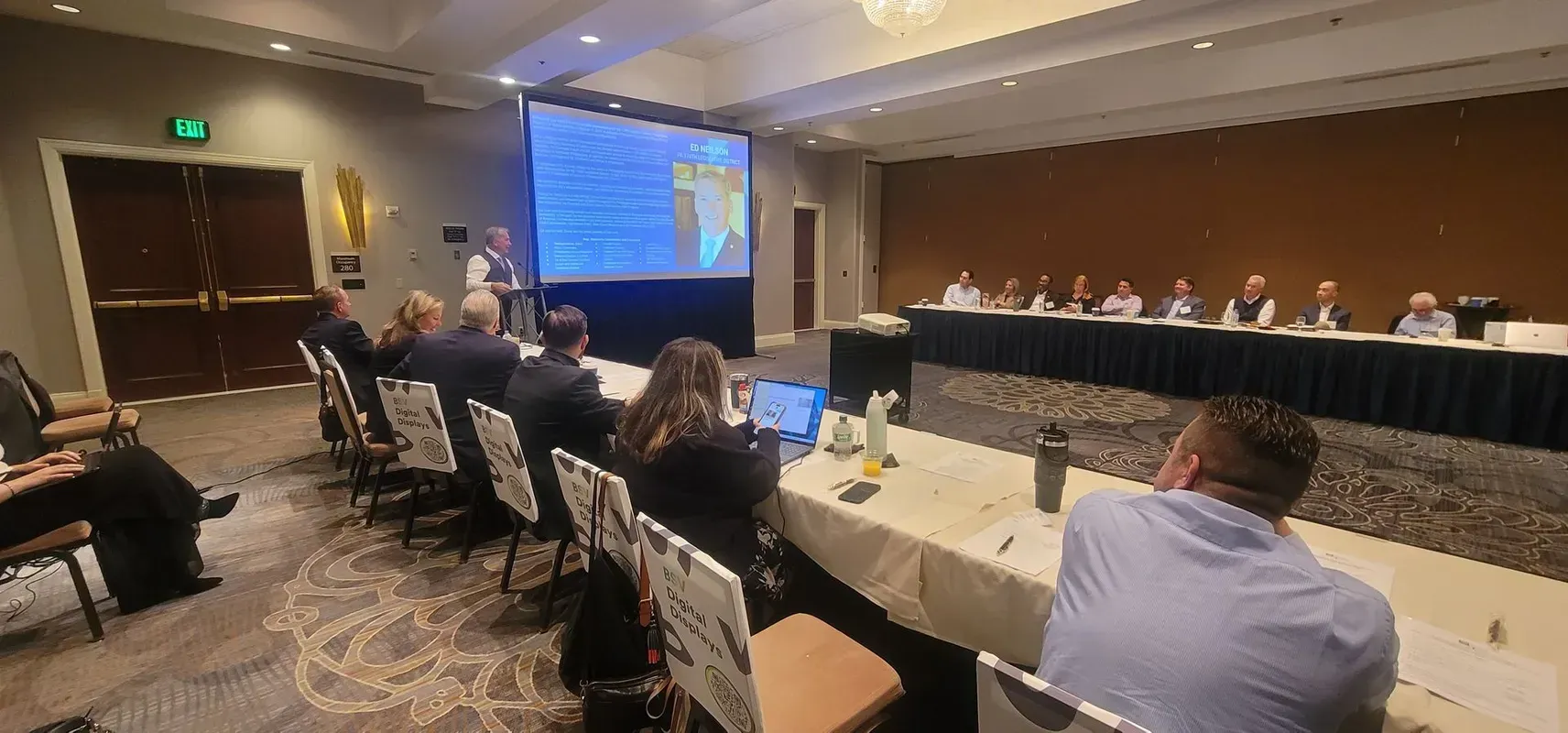 A conference room with presenters and audience members watching a screen with a presentation.