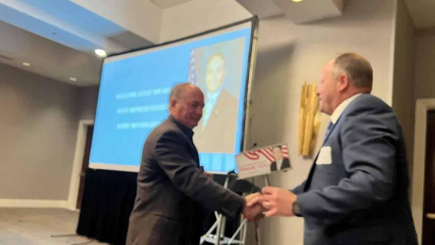 Two men shaking hands in front of a projection screen with a portrait and text. Meeting/conference setting.