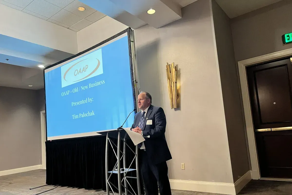 Man in suit giving a presentation at a conference. The screen displays the organization's logo and title.