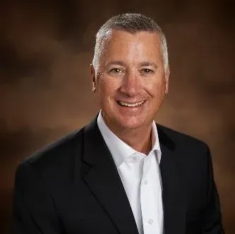 Man with short gray hair, smiling, wearing a dark sweater, collared shirt, and red patterned tie.