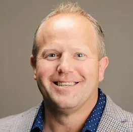 Smiling man in patterned blazer and blue dotted shirt.