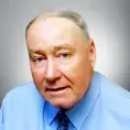 Man in a blue shirt and tie looks forward with a neutral expression against a gray background.