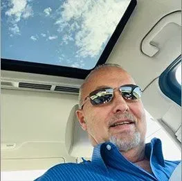 Man in sunglasses smiles from inside a car with a sunroof open, looking at the sky.