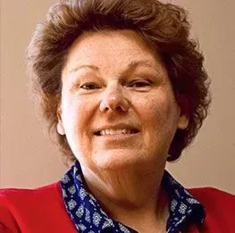 Woman with curly brown hair, smiling, wearing a red jacket over a blue patterned blouse.