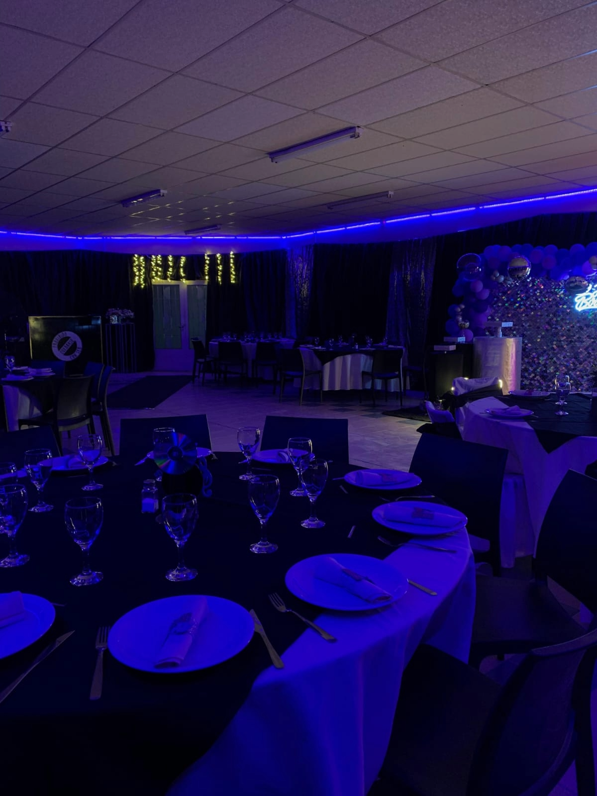 Salón de banquetes tenuemente iluminado, con luces azules y violetas, mesas preparadas y un panel decorativo iluminado en la pared.