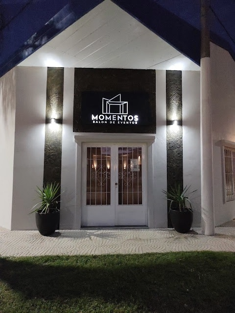 Entrada iluminada de un edificio blanco con un letrero negro, puertas dobles y plantas en macetas.