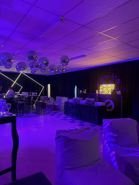 Salón iluminado con luces moradas, con mesas de banquete, sillas y luminarias geométricas en el techo
