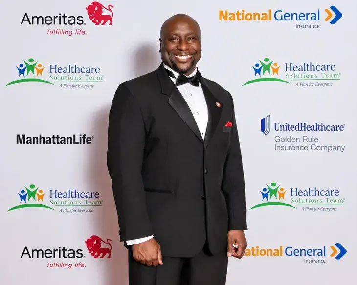 Man in a black tuxedo posing on a step-and-repeat backdrop with sponsor logos