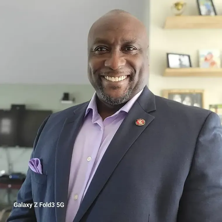 Smiling man in a dark suit and lavender shirt standing indoors in a bright room
