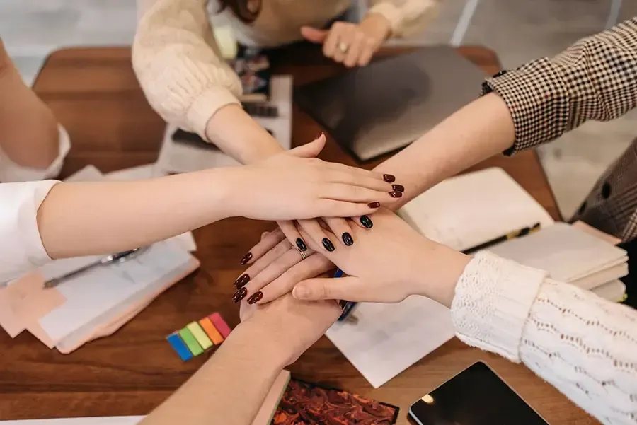 Hands stacked together over a wooden table with papers, notebooks, and colorful markers.