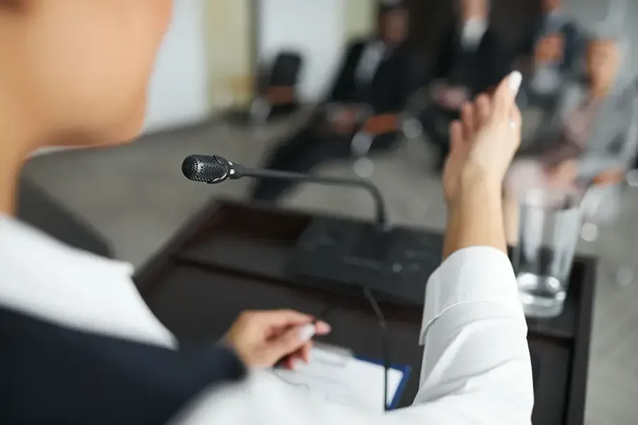 Speaker at a conference podium gestures toward an audience with a microphone in the foreground