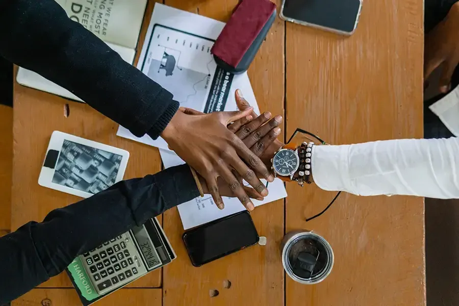 Hands stacked together over a wooden table with papers, laptop, calculator, and coffee cup.