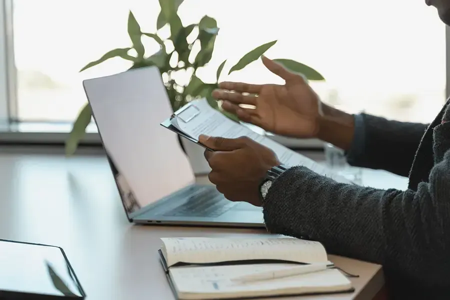 Hands holding a smartphone and gesturing beside a laptop on a desk with an open notebook