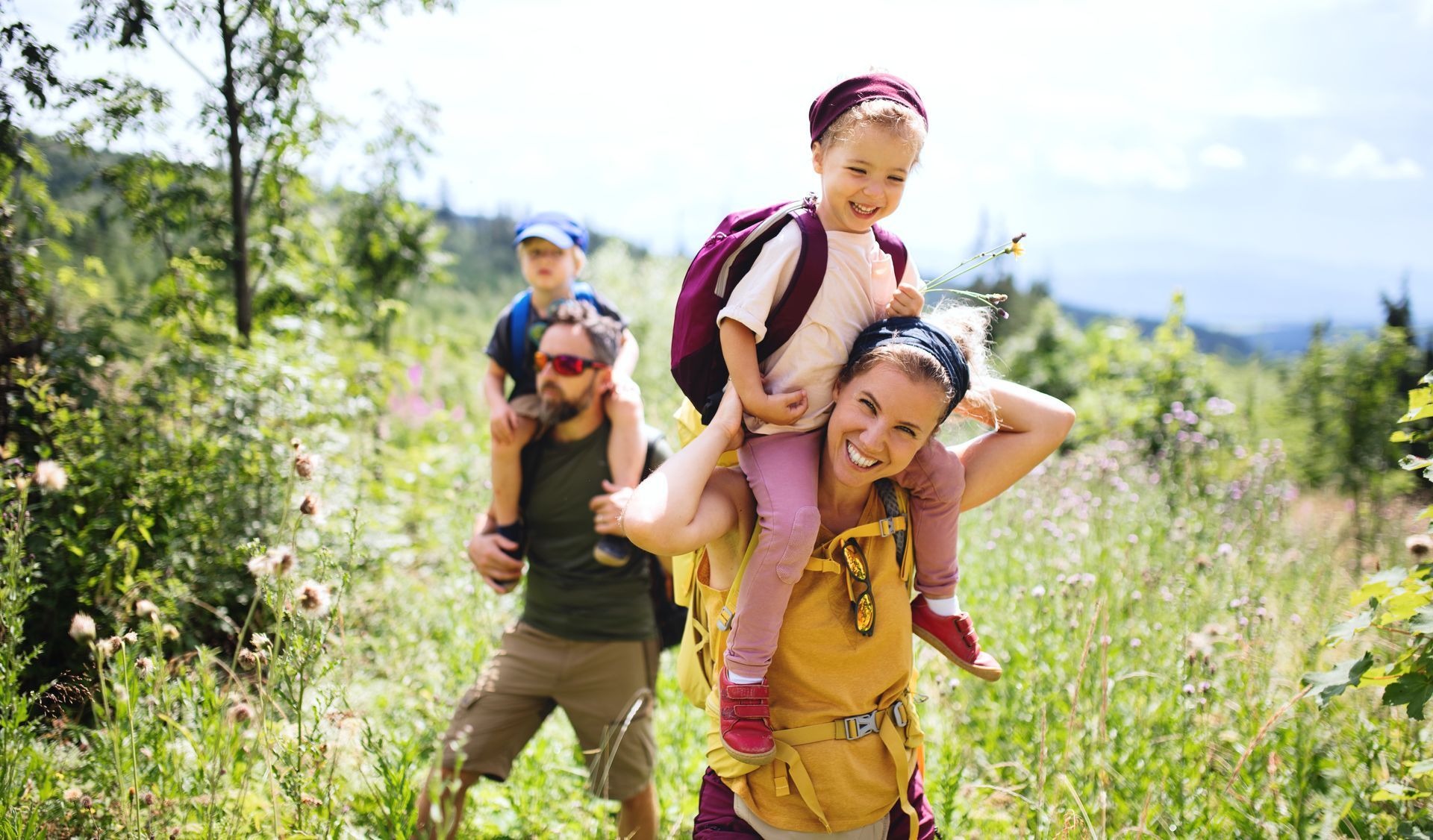 Zwei Erwachsene tragen Kinder auf dem Rücken in einer sonnigen Wiese mit Bäumen und Bergen im Hintergrund.