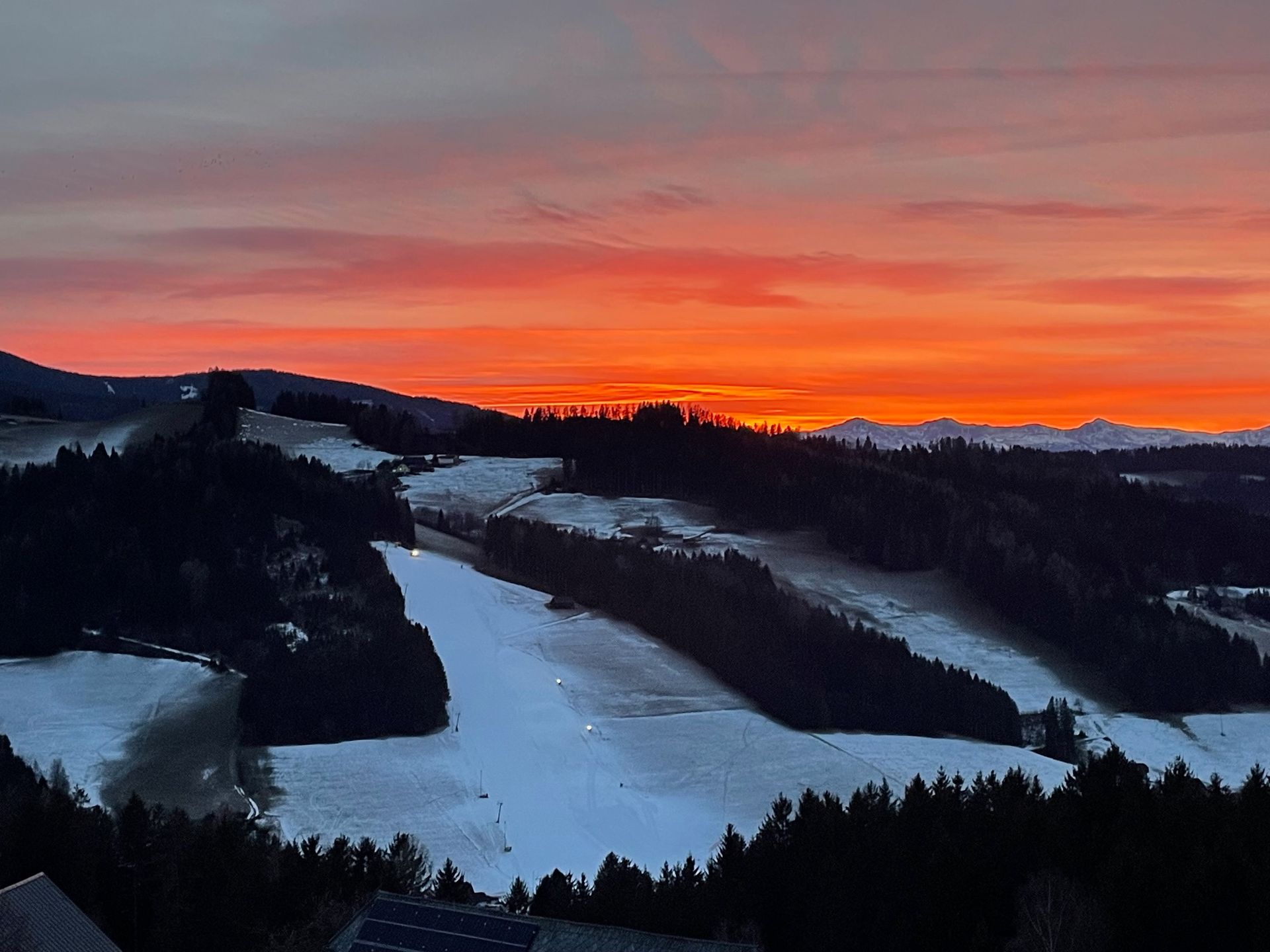 Sonnenuntergang über schneebedeckten Berghängen und dunklen, bewaldeten Hügeln unter einem leuchtend orangefarbenen Himmel
