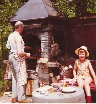 Ein Mann grillt auf einer Terrasse neben einem sitzenden Kind; davor steht ein Tisch mit Getränken und Speisen.