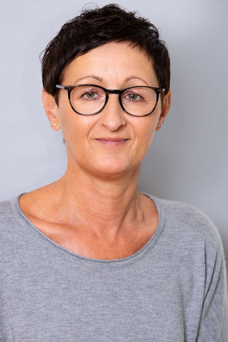 Eine Frau mit Brille und grauem Hemd lächelt in die Kamera.