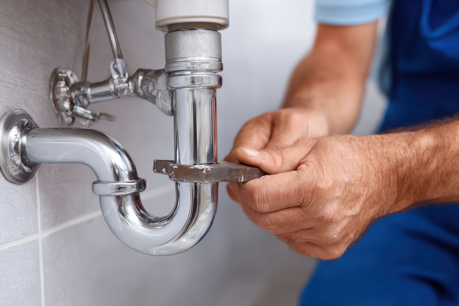 A plumber in blue workwear uses a metal wrench to tighten a chrome P-trap pipe under a sink.