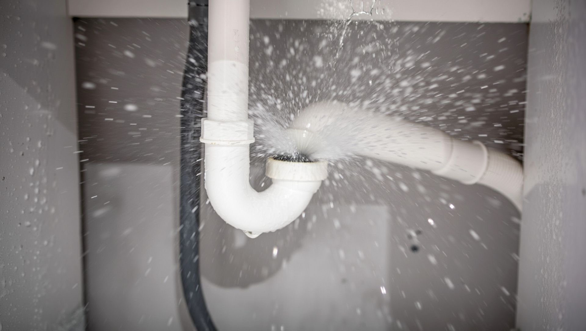 A white P-trap under a sink with water spraying forcefully from a leaking pipe connection.