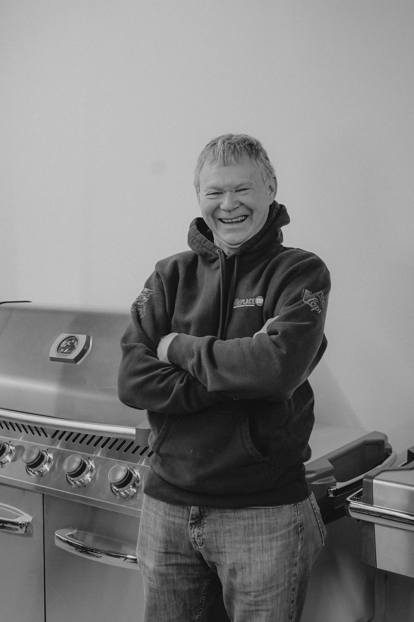 Man in hoodie smiles, arms crossed, beside a stainless steel grill.