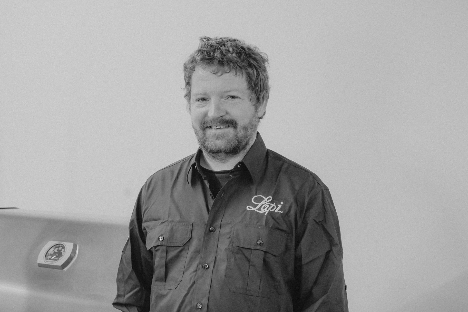 Man with curly hair and a beard, wearing a dark shirt with a logo, standing in front of a stainless steel appliance.