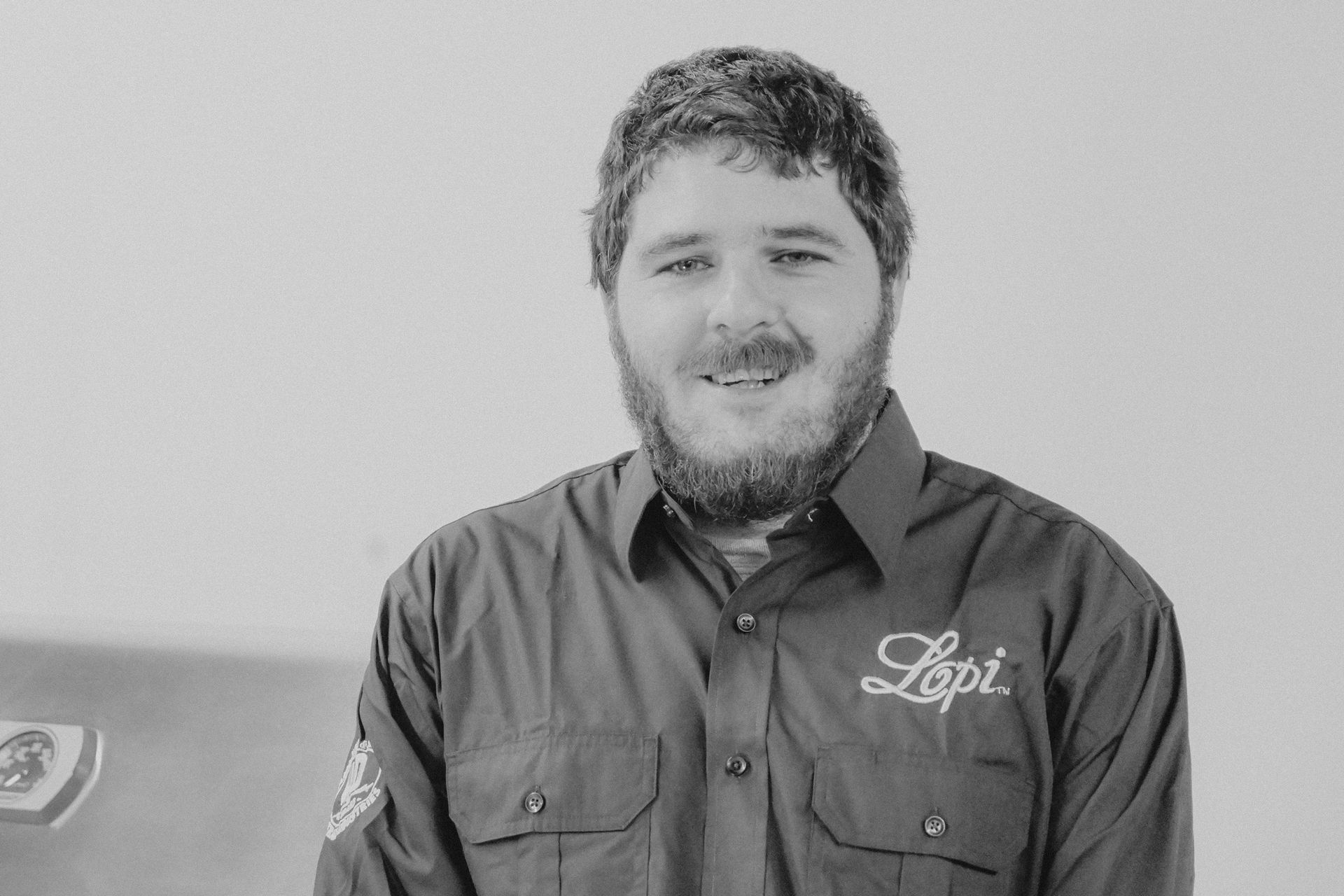 Man in dark shirt with beard, smiling, in front of a light background and a grill.