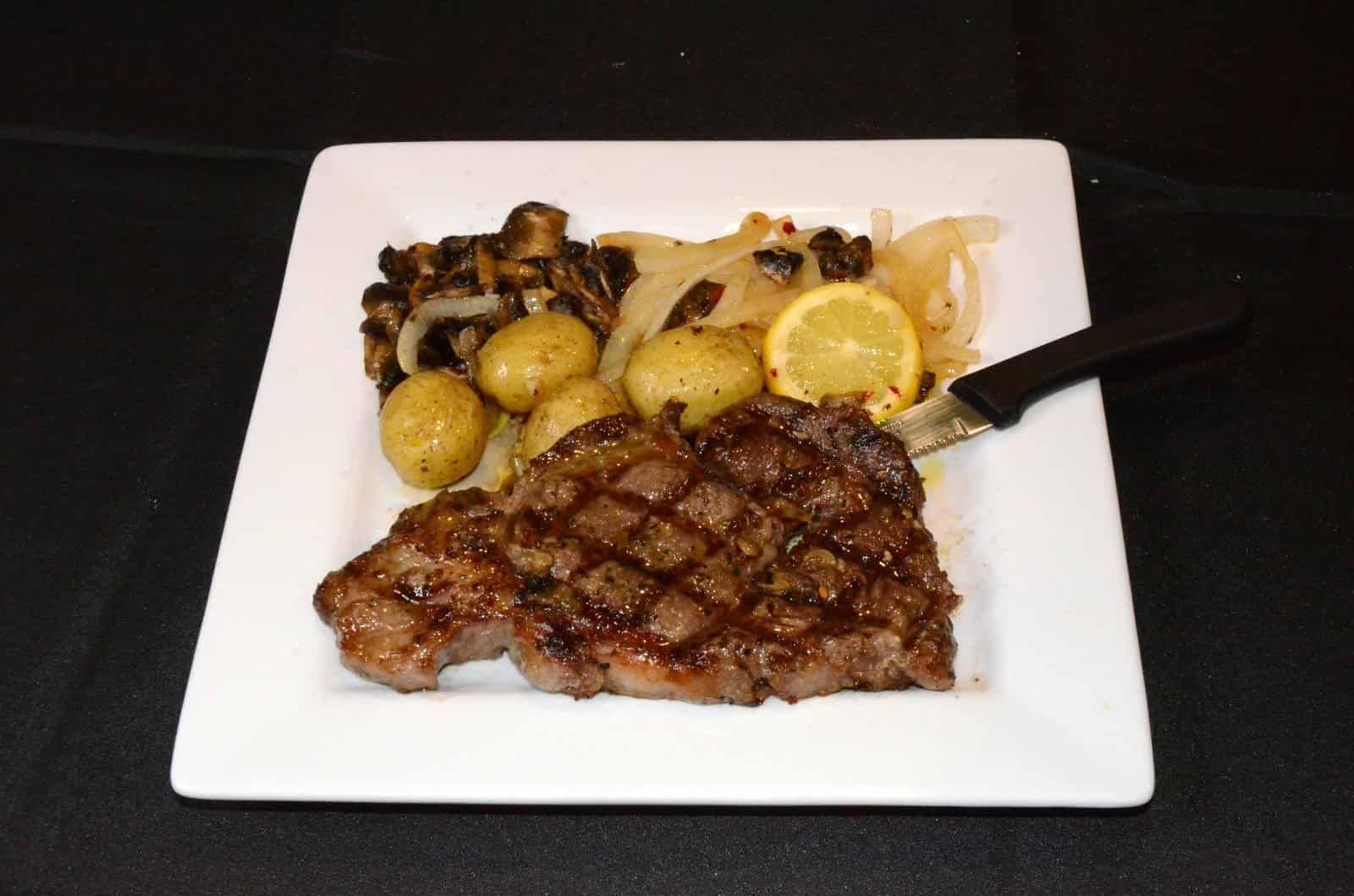 Grilled steak with potatoes, onions, mushrooms, and lemon on a white plate.