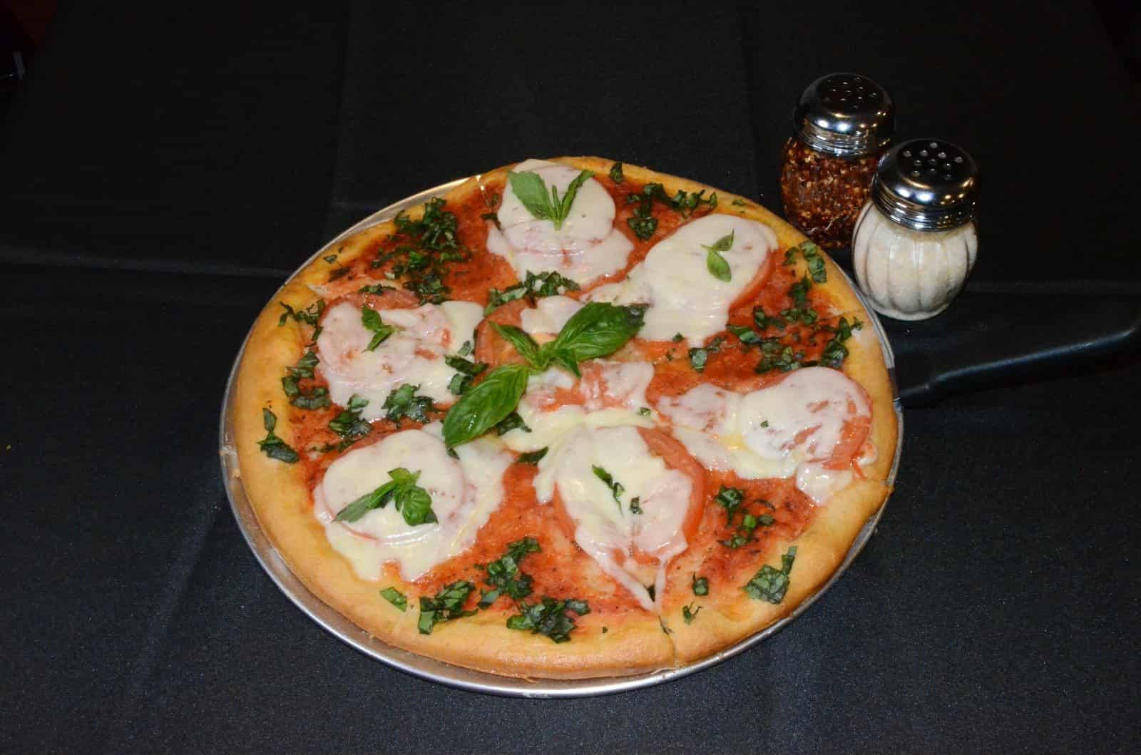 Pizza with tomato, mozzarella, basil on a black background, with pepper and salt shakers.