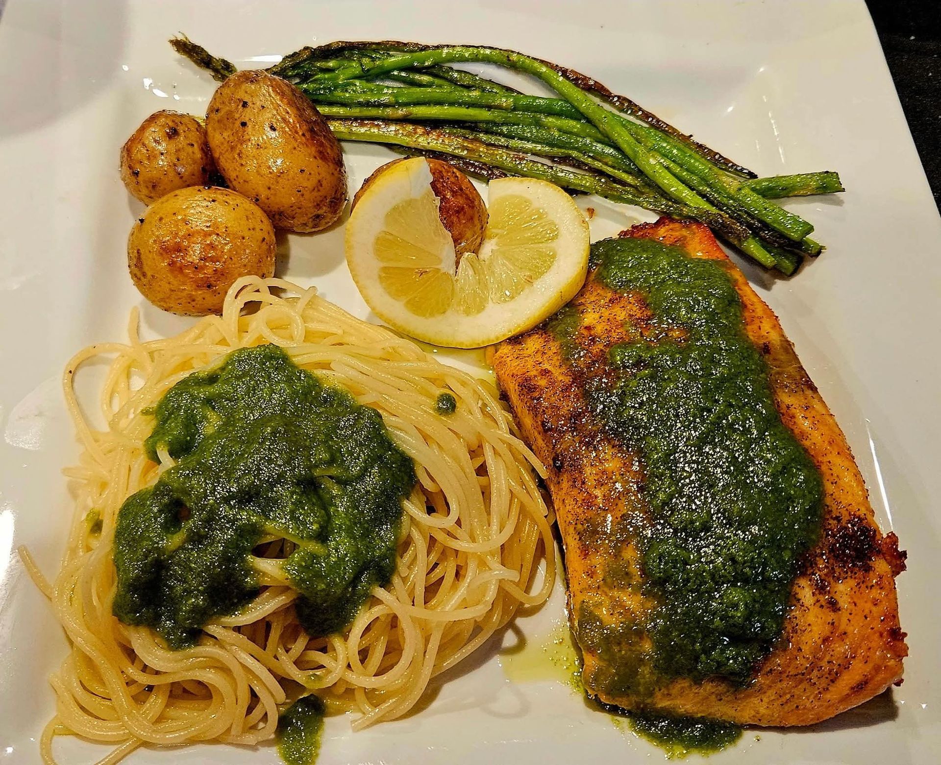 Plate of grilled salmon with pesto, spaghetti, roasted potatoes, and asparagus with lemon.
