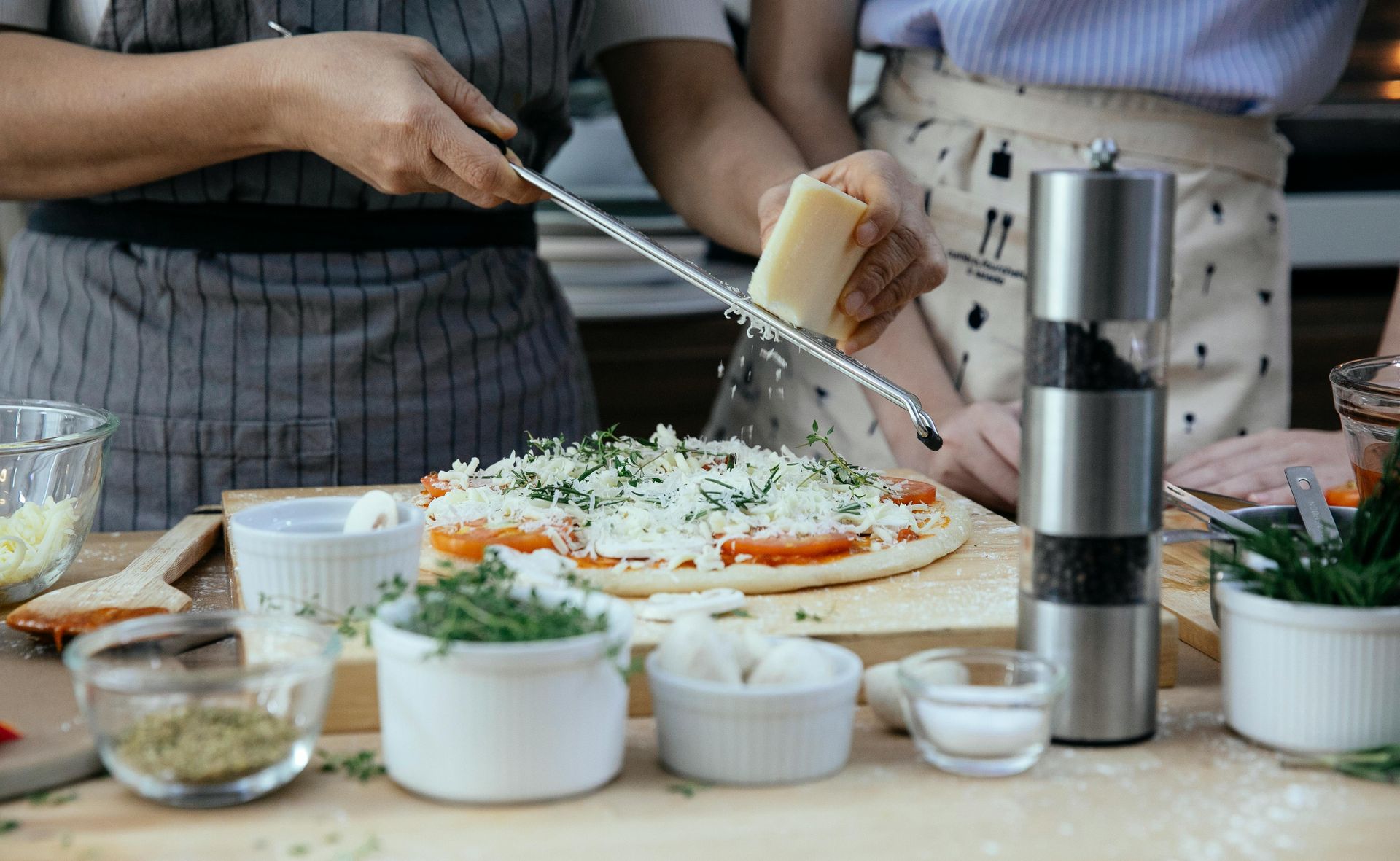 Two people grate cheese onto a pizza, kitchen setting.