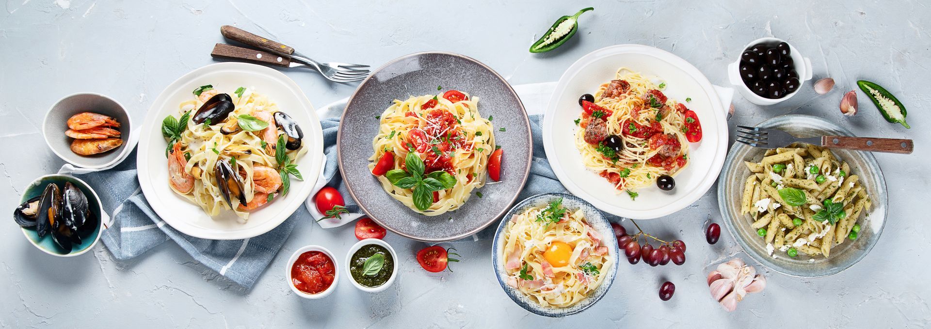 Variety of pasta dishes on a light gray surface, with tomatoes, olives, herbs, and condiments.