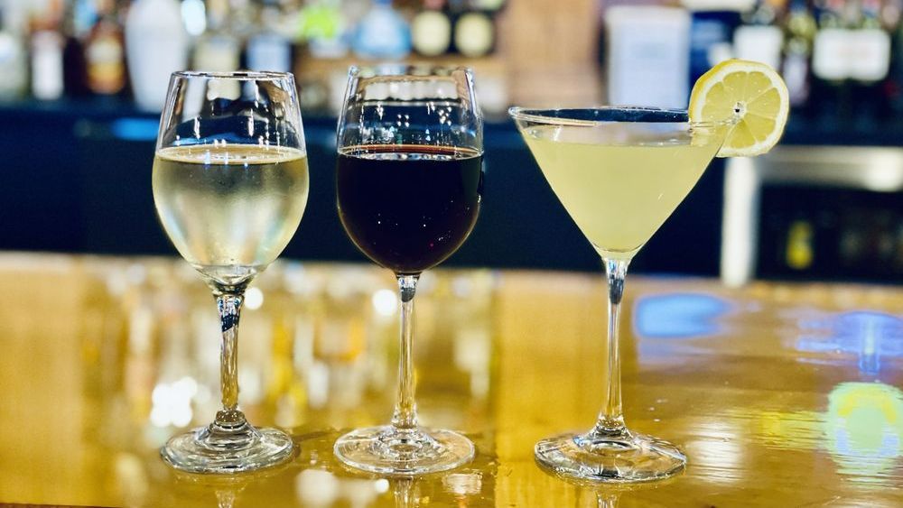Three stemmed glasses on a bar. White wine, red wine, and a cocktail garnished with lemon.